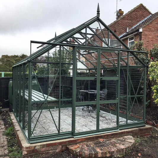 Rhino Greenhouse with dark green metal framing and glass panels is unoccupied, standing on a brick foundation. It is nestled in a garden with surrounding trees and adjacent brick houses.