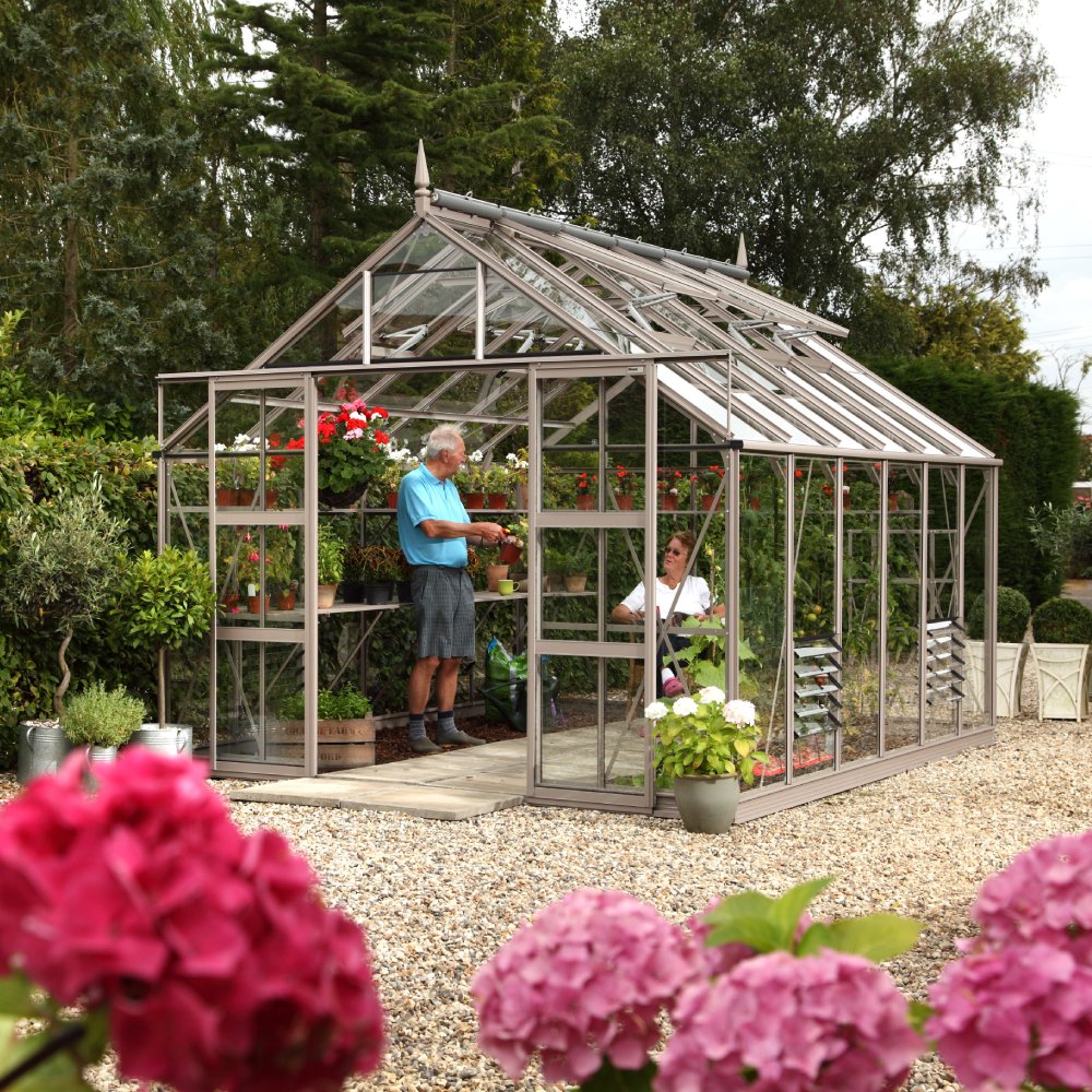 A Rhino Greenhouse filled with potted plants, housing two people interacting inside, is surrounded by a gravel pathway and lush greenery, with pink flowers in the foreground.