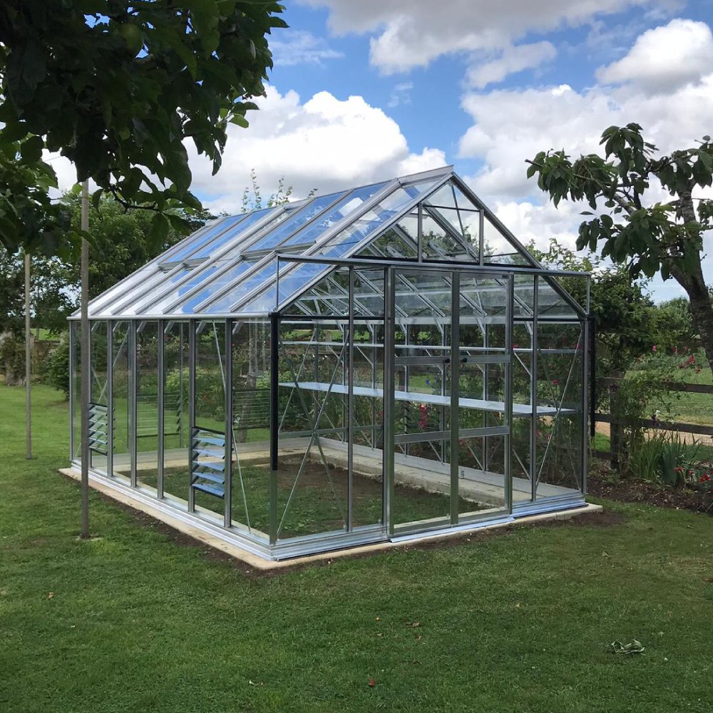 A glass-paneled Rhino Greenhouse stands on a grassy lawn, surrounded by trees and shrubs, under a blue sky with fluffy clouds.