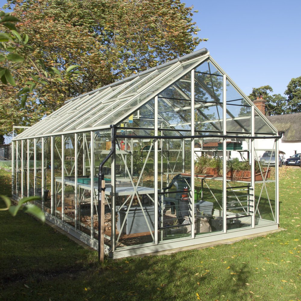 A glass Rhino Greenhouse stands on a grassy lawn, filled with gardening tools and plants. Bathed in sunlight, it is surrounded by trees and a few distant buildings.