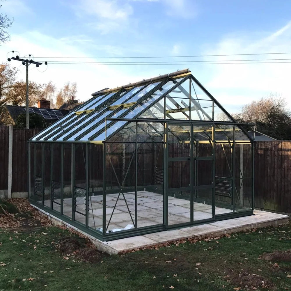A glass and metal Rhino Greenhouse stands on a concrete base, surrounded by grass and a wooden fence, under a blue sky with power lines overhead.