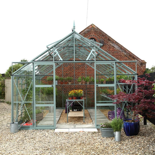 A glass Rhino Greenhouse stands prominently, housing potted plants on tables, in front of a brick building, surrounded by gravel and additional potted plants.