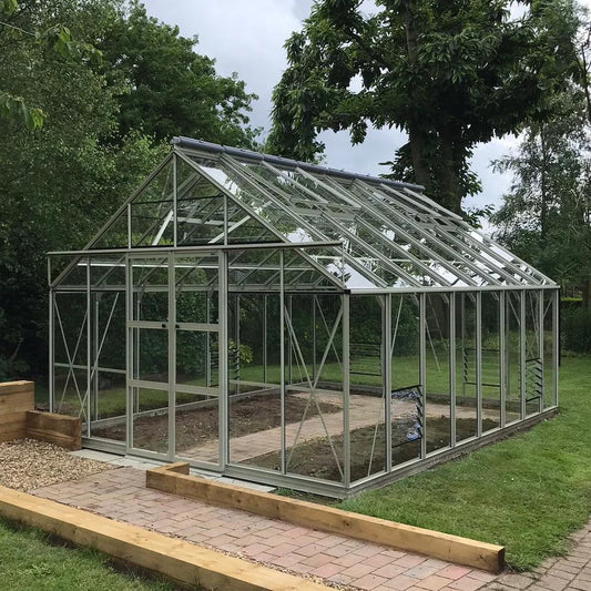 A glass Rhino Greenhouse stands on a paved path surrounded by lush greenery and trees, with a well-manicured lawn extending around the structure.