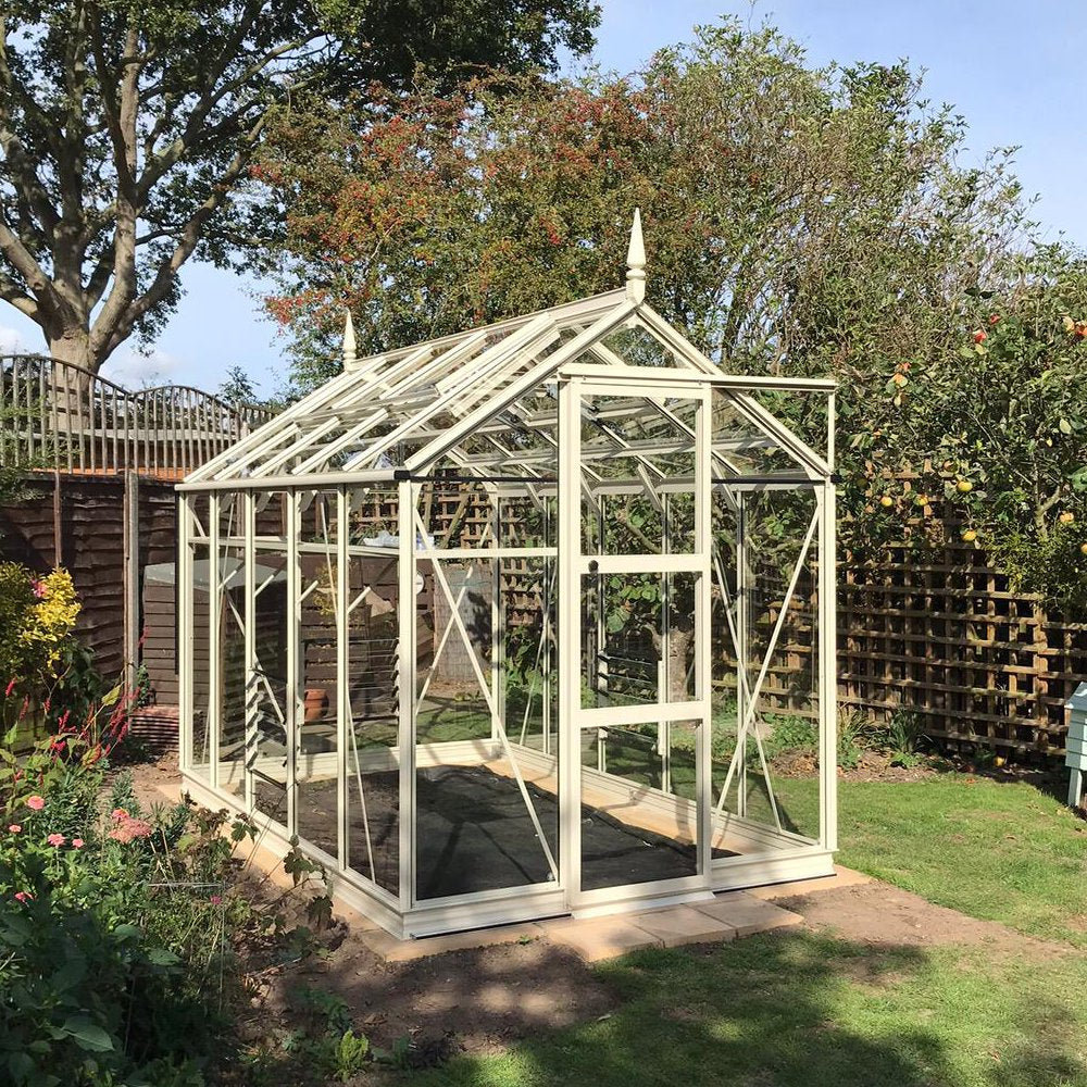 A glass Rhino Greenhouse stands on a garden plot, surrounded by blooming flowers and lush trees. It features a pitched roof with triangular gables and finials, and the sun casts shadows around.