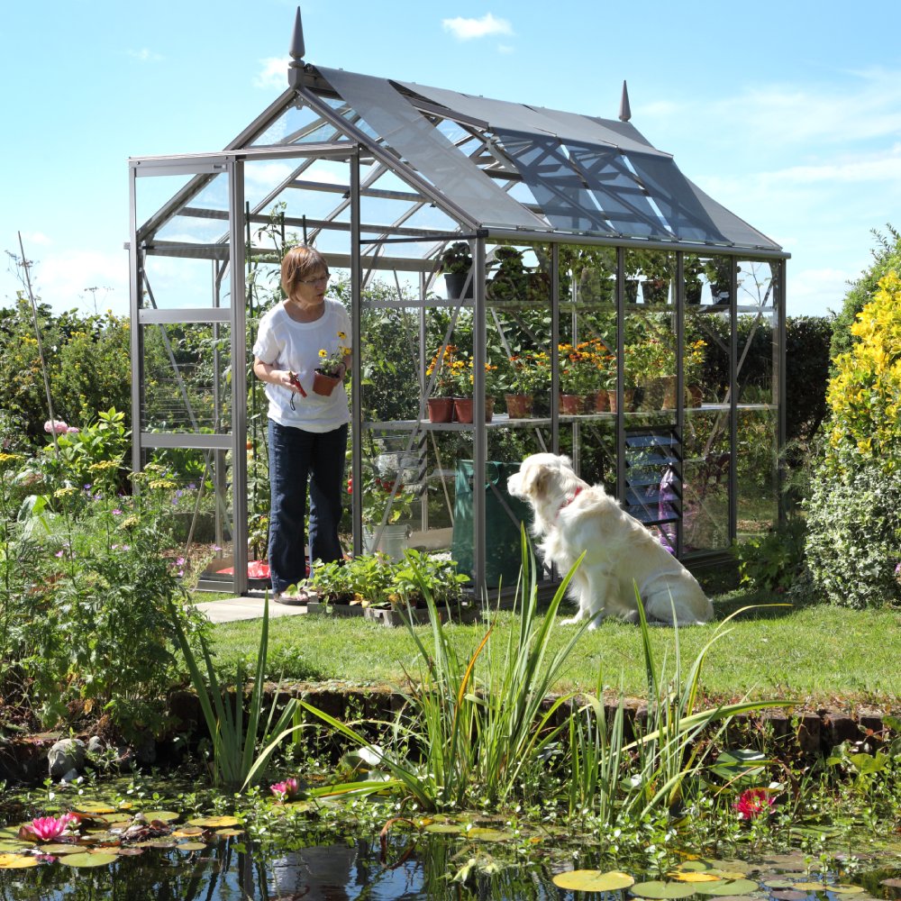 A woman tends plants in a glass-walled Rhino Greenhouse beside a pond, with a dog sitting on the grass. The garden features various flowers and shrubs under a clear blue sky.