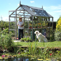 A woman tends plants in a glass-walled Rhino Greenhouse beside a pond, with a dog sitting on the grass. The garden features various flowers and shrubs under a clear blue sky.