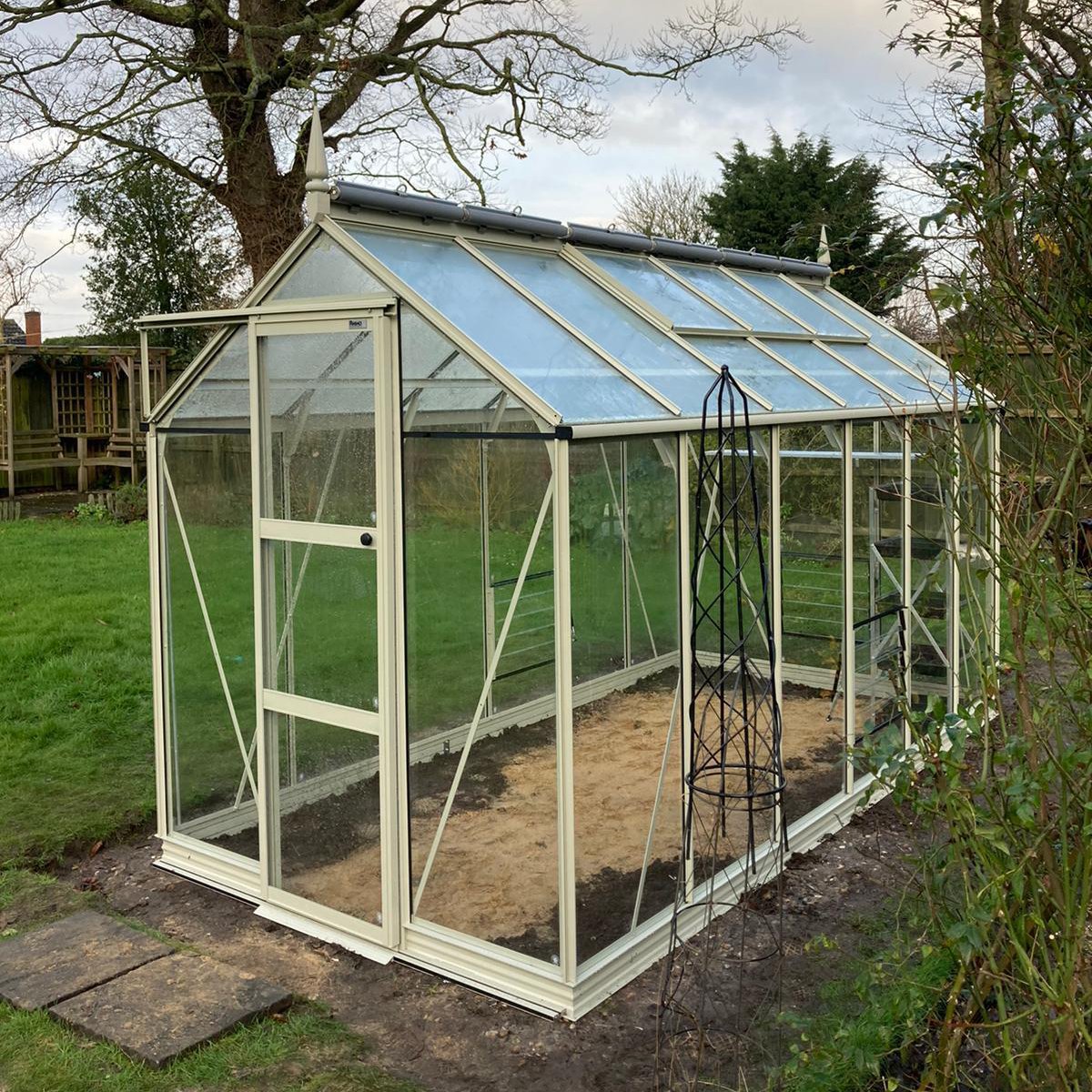 A glass Rhino Greenhouse with a white frame stands on soil, surrounded by grass and trees, under an overcast sky. A metal plant support is positioned nearby.