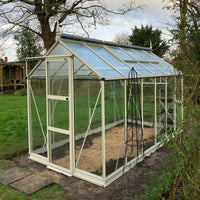 A glass Rhino Greenhouse with a white frame stands on soil, surrounded by grass and trees, under an overcast sky. A metal plant support is positioned nearby.