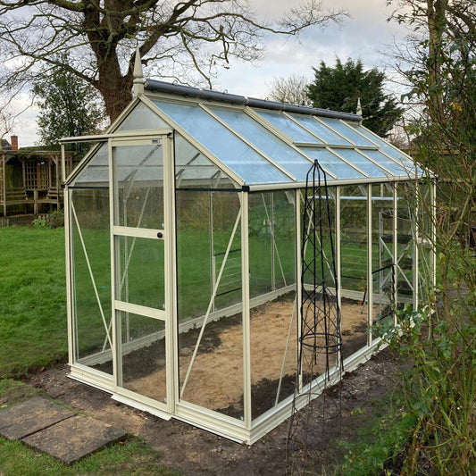 A glass Rhino Greenhouse with a white frame stands on soil, surrounded by grass and trees, under an overcast sky. A metal plant support is positioned nearby.