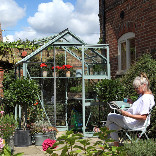 Rhino Greenhouse with potted plants inside, surrounded by brick walls and foliage. A person sits nearby in a chair, reading a newspaper and holding a blue cup, under a sunny blue sky.
