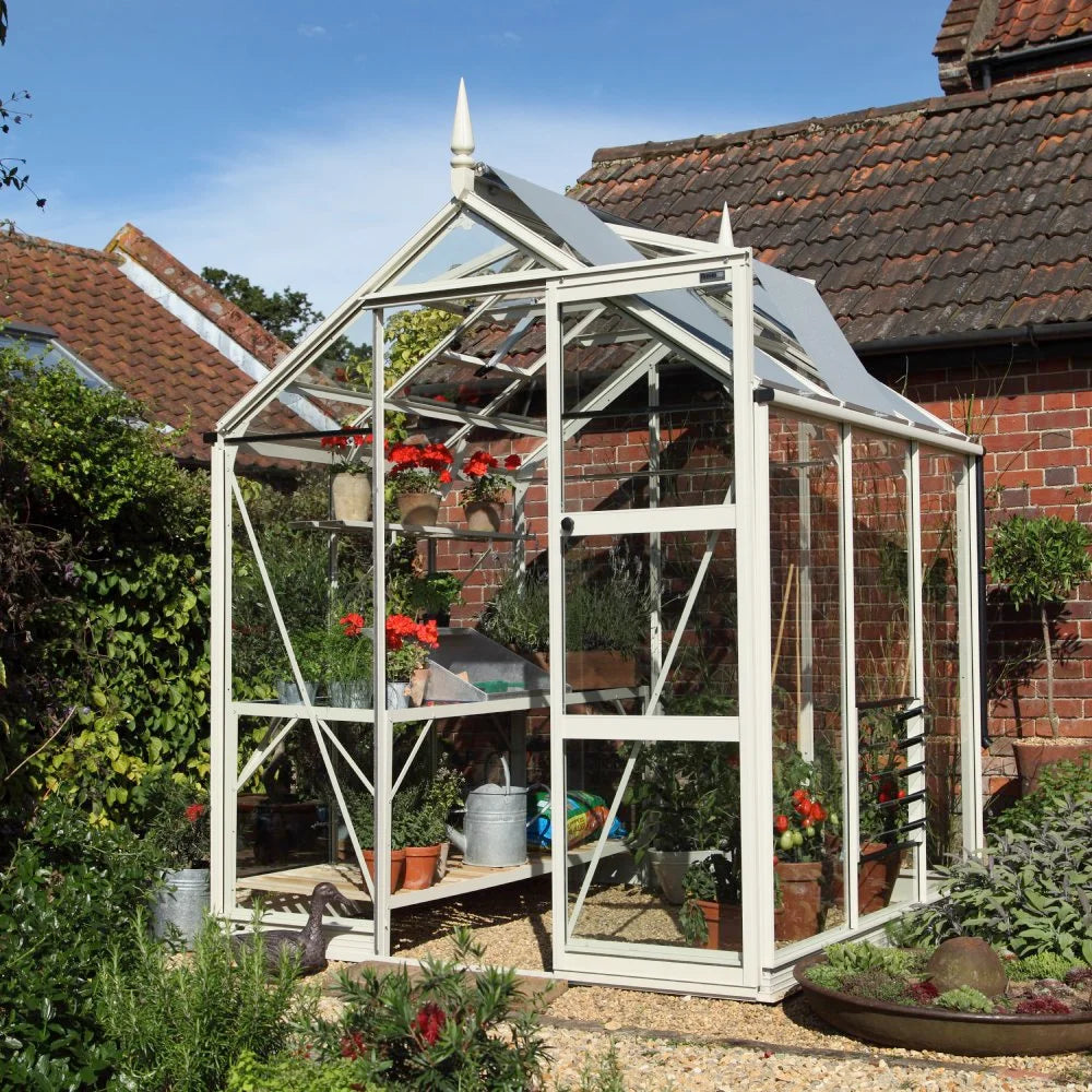 A white-framed Rhino Greenhouse stands in a garden, housing potted red flowers and gardening tools, adjacent to a brick wall with a tiled roof.