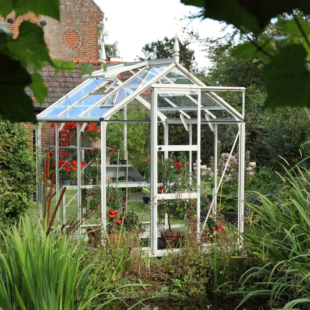 A glass Rhino Greenhouse houses vibrant red flowers and potted plants, surrounded by lush greenery, with a brick building partially visible in the background.
