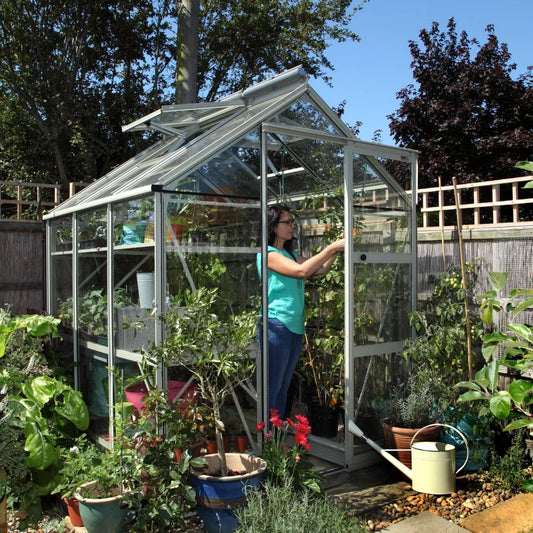 A woman tends plants inside a glass Rhino Greenhouse surrounded by potted greenery. Sunlight filters through, illuminating the thriving plants. A yellow watering can sits outside on a stone path.