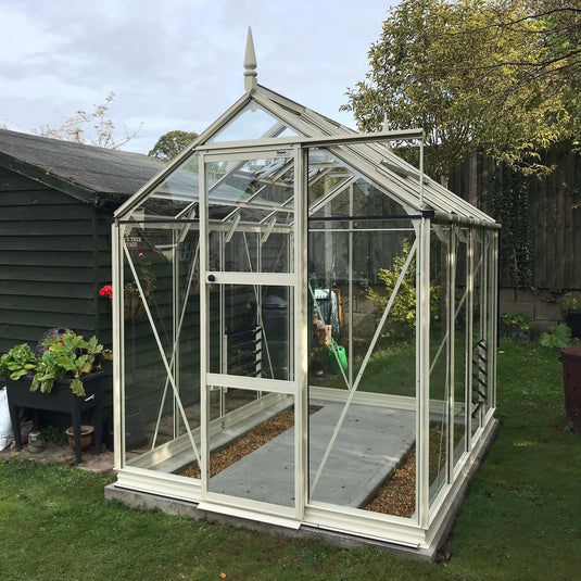 A glass Rhino Greenhouse stands on a lawn, featuring a white frame with a pointed roof. It's adjacent to a wooden shed, surrounded by greenery and trees.
