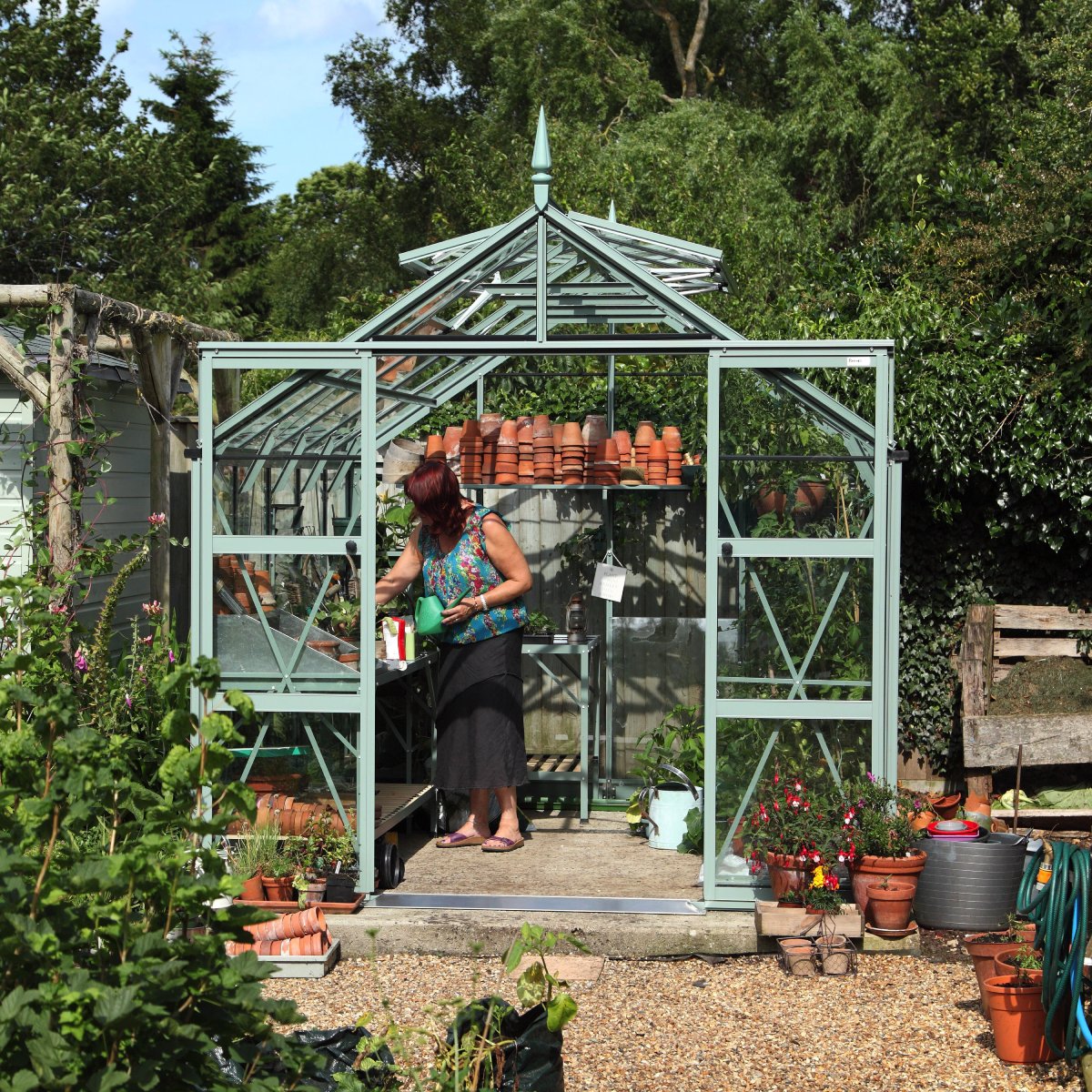 A woman waters plants inside a green metal-framed Rhino Greenhouse filled with terracotta pots and gardening tools. The Rhino Greenhouse is surrounded by lush greenery and garden accessories, under a clear sky.