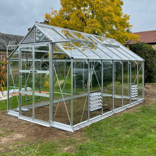 A glass and metal Rhino Greenhouse stands on grass, featuring vented panels, under an overcast sky with autumn trees and nearby rooftops.