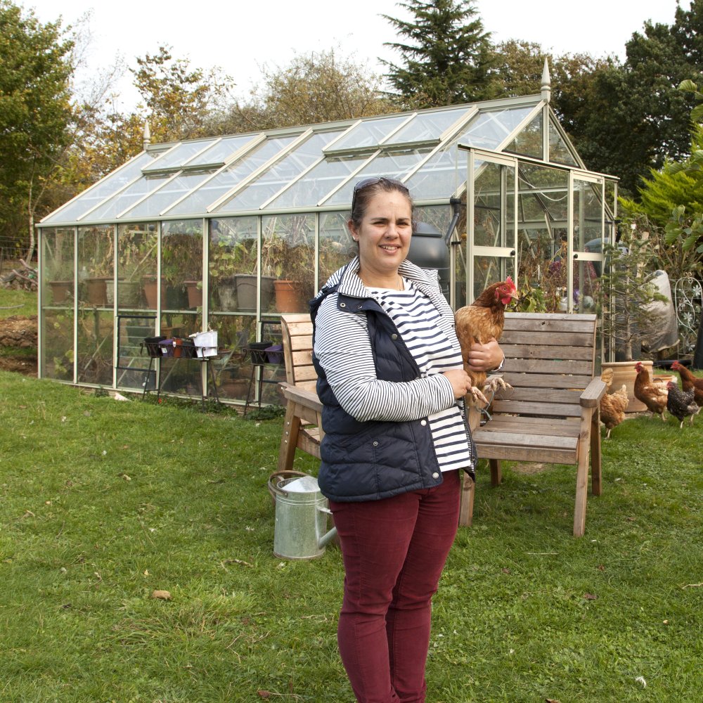 A person holds a chicken while standing in front of a glass Rhino Greenhouse filled with plants. Nearby, a wooden bench, watering can, and additional chickens complete the garden setting.