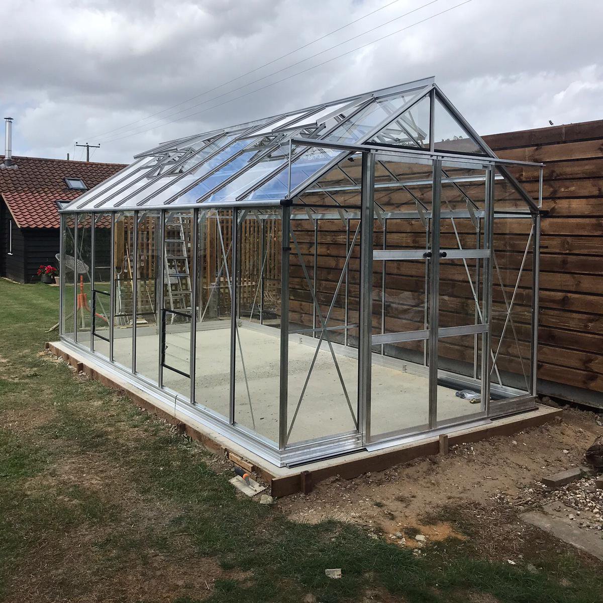 A glass Rhino Greenhouse with metal frame stands on a concrete base, next to a wooden fence. A red-roofed building and a ladder are visible in the grassy yard.