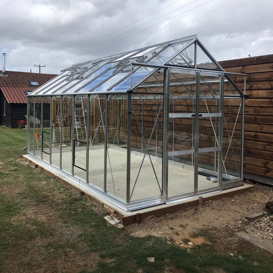 A glass Rhino Greenhouse with metal frame stands on a concrete base, next to a wooden fence. A red-roofed building and a ladder are visible in the grassy yard.