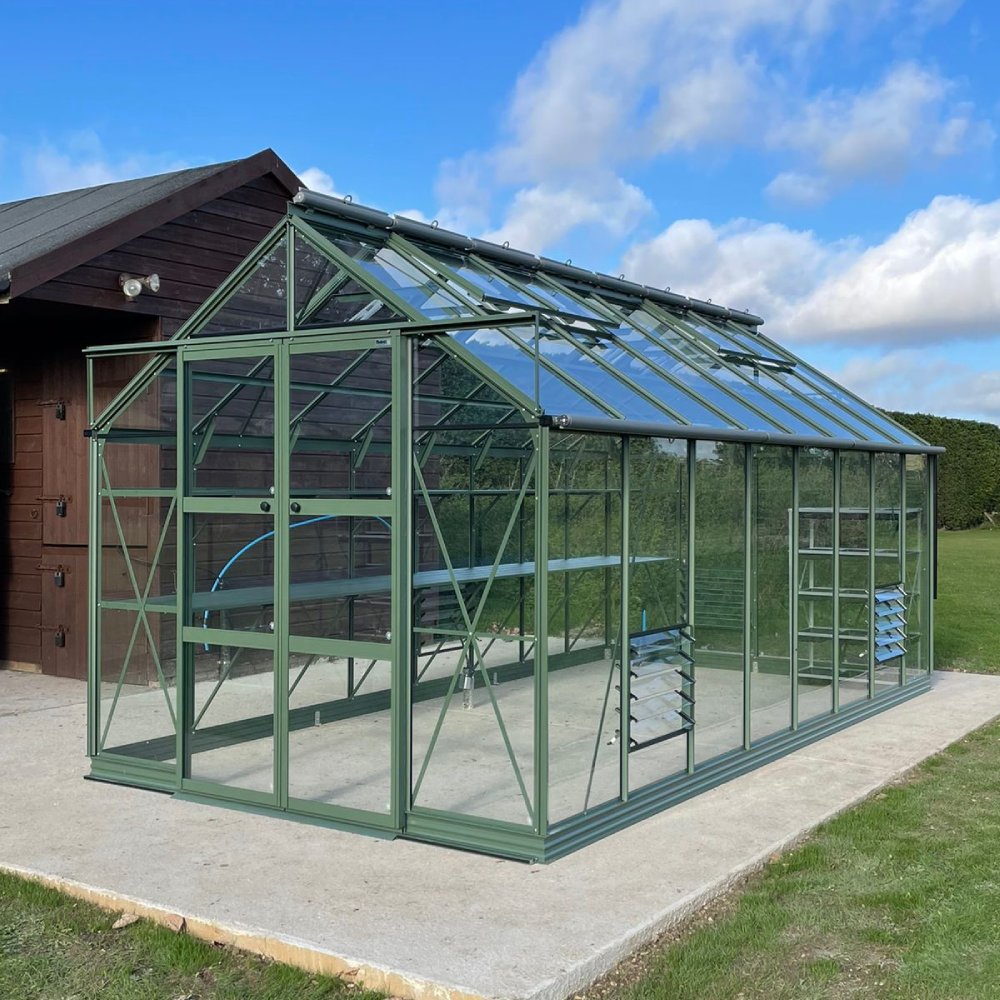 A green metal-framed Rhino Greenhouse stands on a concrete slab, with large glass panels and a door. It's adjacent to a wooden building, surrounded by grass under a blue, cloudy sky.