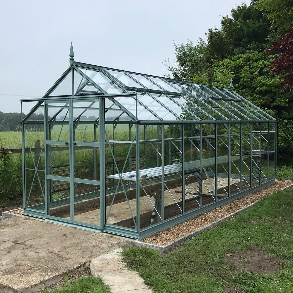 A large glass Rhino Greenhouse stands on a gravel foundation with a metal frame, set amidst a rural garden with lush greenery and trees in the background.