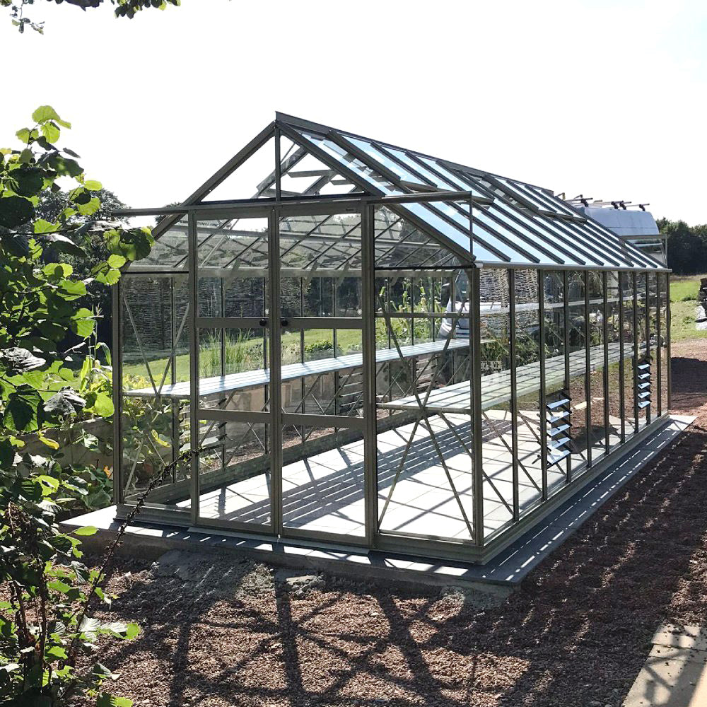 A glass Rhino Greenhouse with open doors, positioned on a gravel path, surrounded by trees and grass, reflecting sunlight.