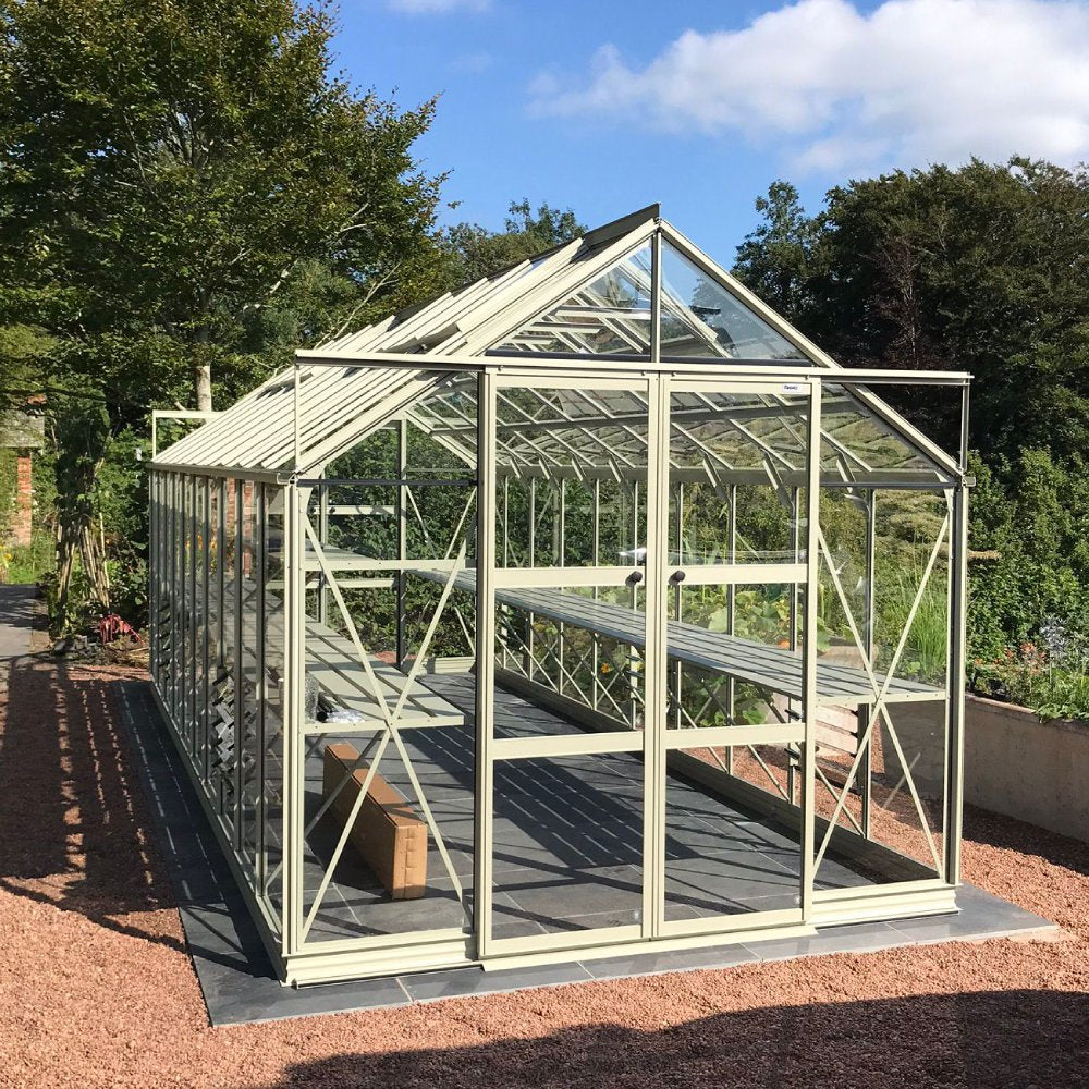 A glass Rhino Greenhouse with a metal frame stands on a gravel path, surrounded by trees and greenery, containing shelves and a cardboard box inside.