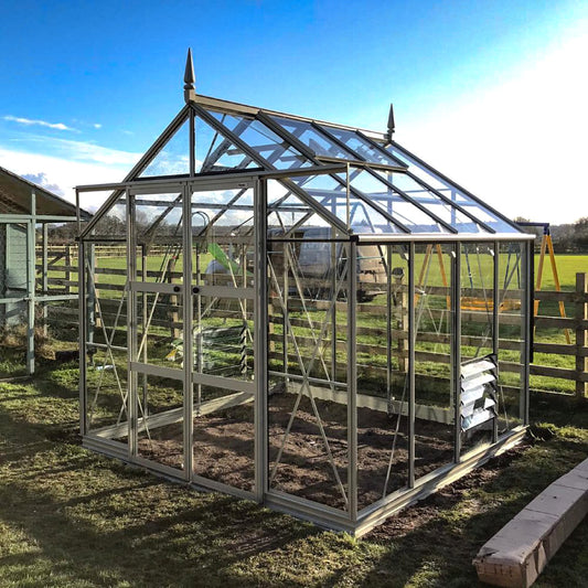 A glass Rhino Greenhouse with a peaked roof and metallic frame stands on a grassy area, surrounded by wooden fencing, under a clear blue sky.