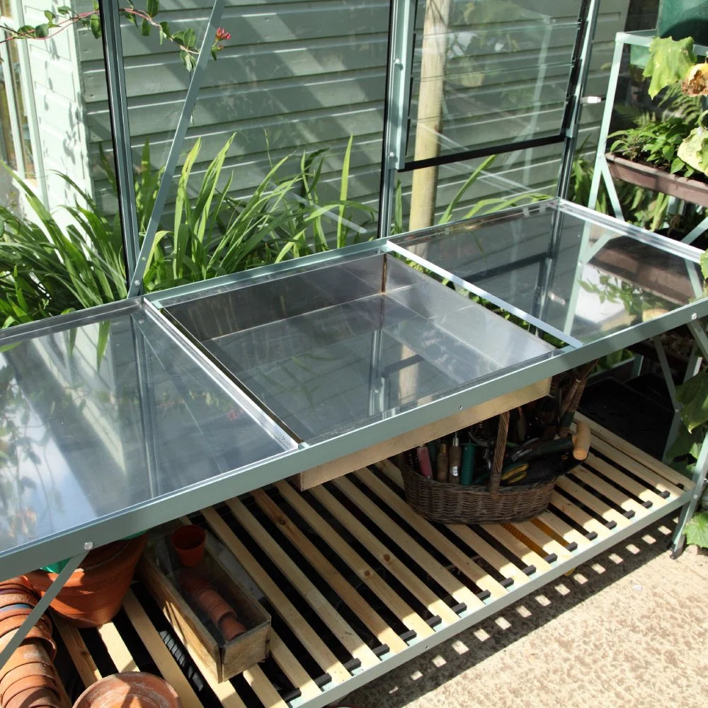 A metal-framed potting bench stands in a Rhino Greenhouse, featuring clear panels and a slatted shelf with gardening tools and terracotta pots stored beneath. Lush green plants surround the area.