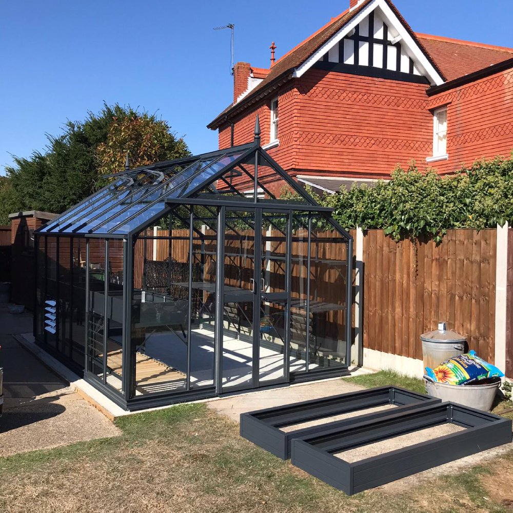 A glass Rhino Greenhouse stands in a backyard next to a red-brick house, surrounded by wooden fencing and gardening supplies such as soil bags and planter frames on the grass.
