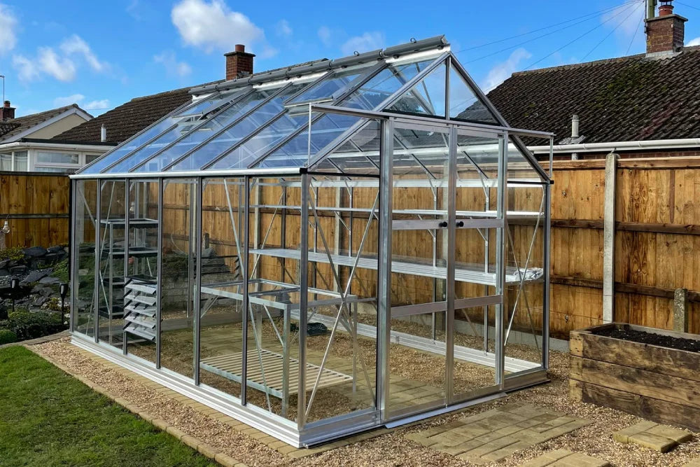 A glass and aluminum Rhino Greenhouse stands in a backyard, with clear panels and metal framing, surrounded by a lawn and wooden fence, under a blue sky.