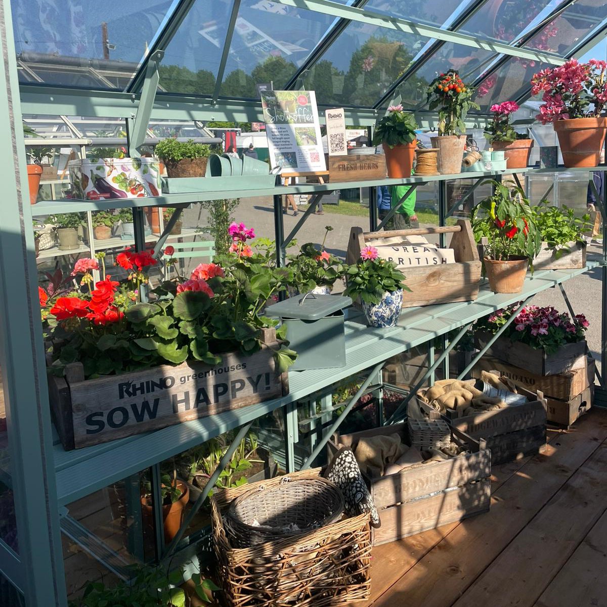 Shelves displaying potted plants and gardening tools in a Rhino Greenhouse. Sunlight filters through the glass roof. A sign reads Rhino Rhino Greenhouses SOW HAPPY!! with other smaller signs nearby.