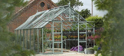 A glass Rhino Greenhouse stands with open doors, surrounded by plants and gardening supplies, next to a brick building and lush greenery.