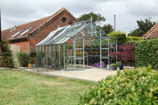 A glass Rhino Greenhouse stands on a gravel area, filled with potted plants. It is situated next to a brick building surrounded by greenery and a wooden fence.