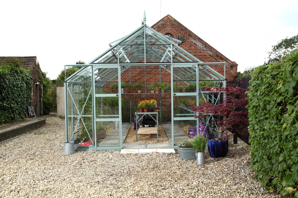 A glass Rhino Greenhouse stands on a gravel pathway, filled with plants and gardening supplies. It's surrounded by lush greenery and a brick structure in the background, creating a cozy garden setting.