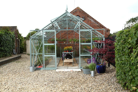 A glass Rhino Greenhouse stands on a gravel pathway, filled with plants and gardening supplies. It's surrounded by lush greenery and a brick structure in the background, creating a cozy garden setting.