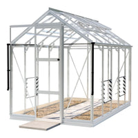 A white-framed Rhino Greenhouse with a hinged door stands on a stone pathway, featuring vented sides and a transparent roof in a spacious outdoor setting.