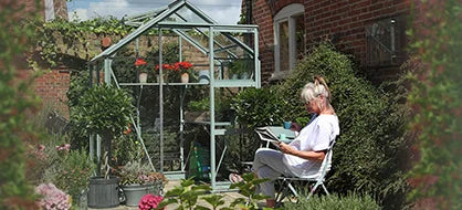 A woman sits reading on a white chair beside a glass Rhino Greenhouse filled with plants. The setting is a brick-walled garden with various potted plants and flowers around.