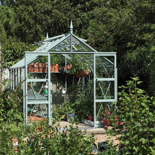 A glass Rhino Greenhouse stands surrounded by lush greenery, containing potted plants and gardening tools, with terracotta pots lined up inside. It's set in a garden with trees in the background.