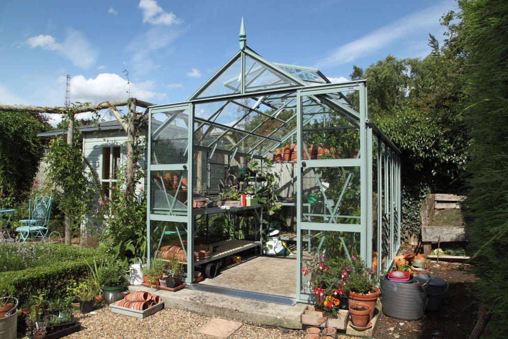 A glass Rhino Greenhouse stands open with potted plants and gardening tools inside, surrounded by lush greenery and outdoor furniture on a sunny day.