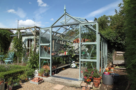 A glass Rhino Greenhouse stands open with potted plants and gardening tools inside, surrounded by lush greenery and outdoor furniture on a sunny day.
