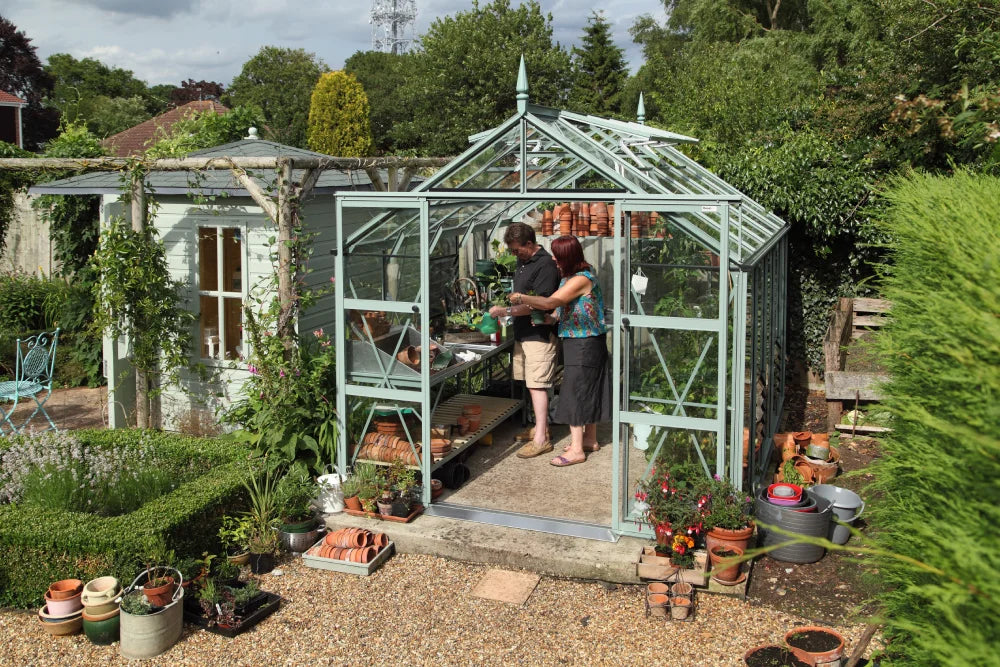 A green metal Rhino Greenhouse houses two people arranging plants and pots. Surrounding hedges and trees frame a garden setting with scattered gardening tools and accessories.