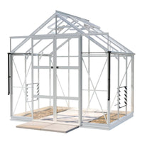 A white-framed Rhino Greenhouse stands with open double doors, showcasing clear panels and roof vents, positioned on a stone-tiled base in a simple, isolated setting.