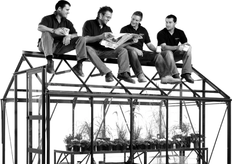 Four people, sitting on a Rhino Greenhouse roof, are engaged in conversation. One holds a newspaper, while others hold mugs. Inside the Rhino Greenhouse, plants are organized on shelves.