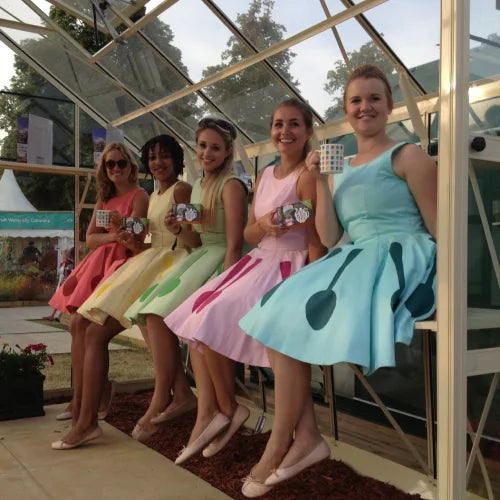 Five women, wearing colorful dresses with polka dots, sit on a wooden bench inside a Rhino Greenhouse. Each holds a small container. Trees and garden structures are visible outside.