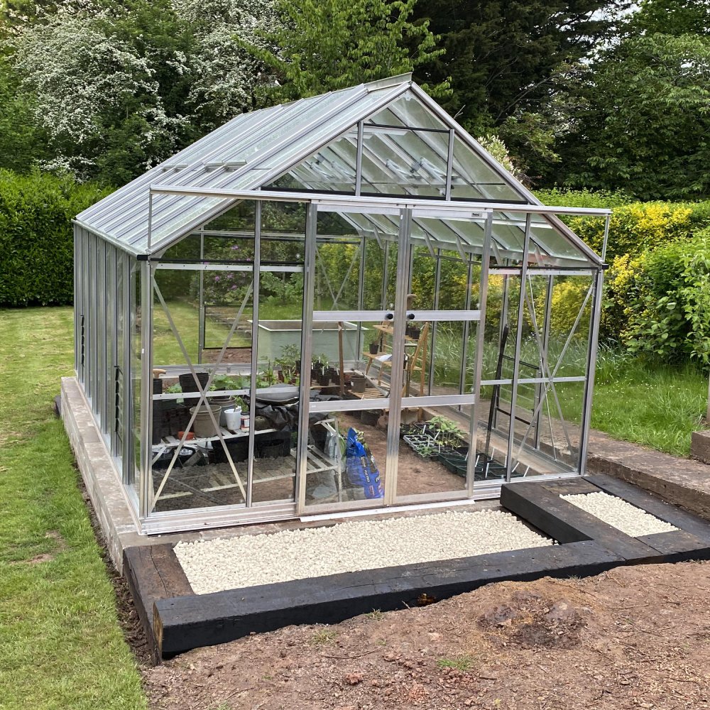 A large glass Rhino Greenhouse stands on a landscaped garden, filled with various potted plants and gardening tools, surrounded by lush greenery and bordered by gravel and wooden beams.