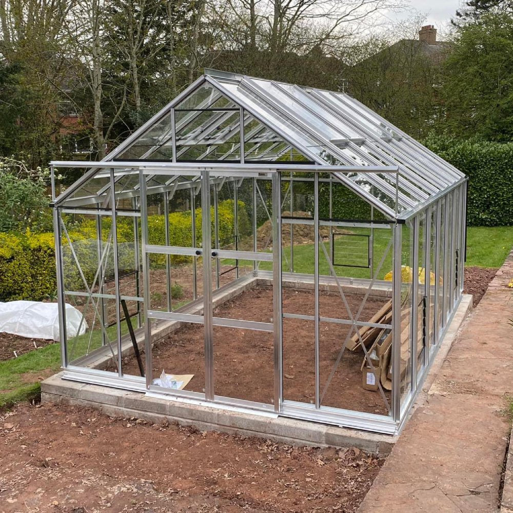 A glass and metal Rhino Greenhouse stands assembled, empty with opened doors, surrounded by a garden with trees and bushes, situated on a dirt foundation.