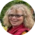 A person with curly hair and glasses smiles, wearing a red scarf. The background features green foliage.