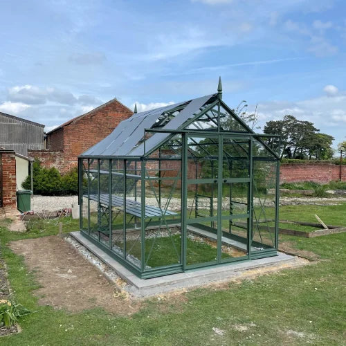 A green metal-framed Rhino Greenhouse stands on a concrete base, surrounded by grass and near brick buildings under a partly cloudy sky.