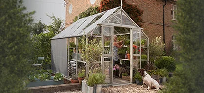 A Rhino Greenhouse stands in a lush garden, with a person tending to plants inside. Pots and a dog sit nearby, surrounded by trimmed hedges and a brick house in the background.